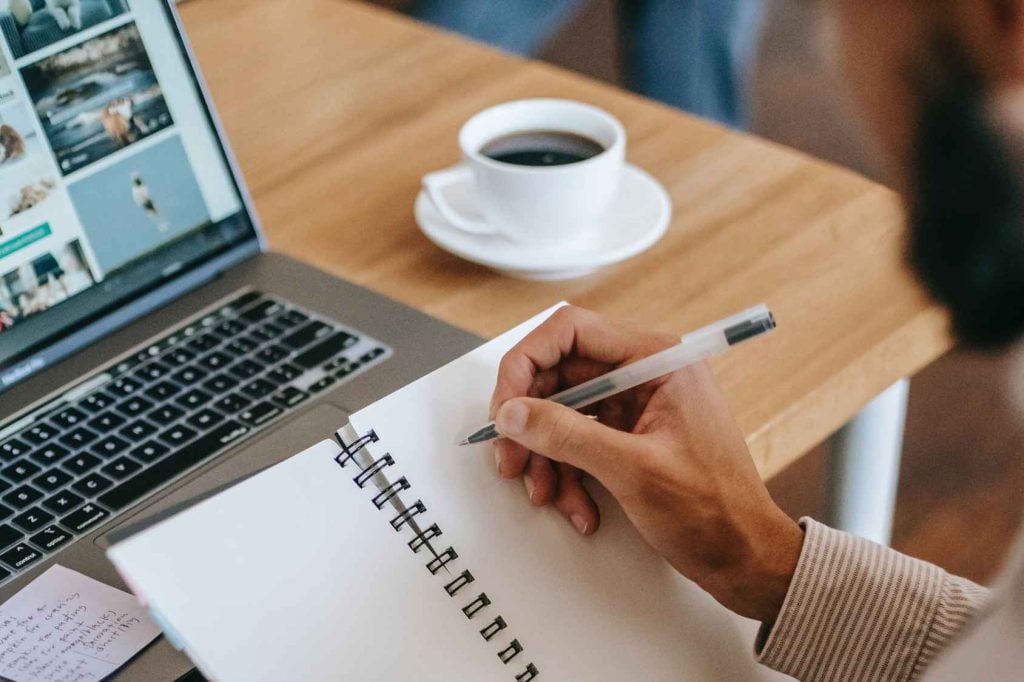 man writing in notebook in front of laptop